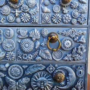 Puede incluir: Una caja de madera azul con tres cajones decorados con varios adornos en 3D, incluyendo flores, hojas, mariposas, un sol, una luna y una esfera de reloj. La caja tiene tiradores de color dorado.