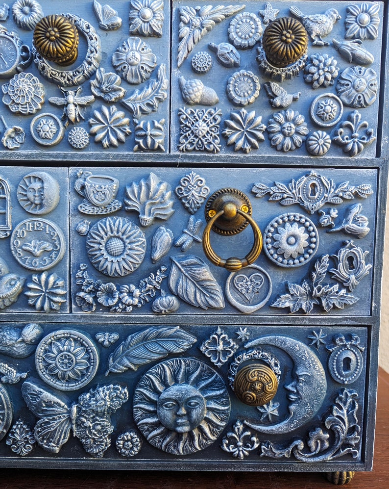 Puede incluir: Una caja de madera azul con tres cajones decorados con varios adornos en 3D, incluyendo flores, hojas, mariposas, un sol, una luna y una esfera de reloj. La caja tiene tiradores de color dorado.