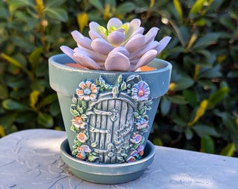 Puede incluir: Una pequeña maceta de cerámica de color verde azulado con una puerta decorativa y detalles florales. La maceta está llena de una planta suculenta morada.