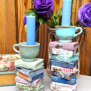 May include: Two teal blue teacup candle holders with blue candles, each sitting atop a stack of vintage-looking books. The books have various colors and patterns, including pink, blue, green, and white. One stack of books has a keyhole and a key on the top book. The other stack of books has the text "ventorum" on the top book.