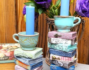 May include: Two teal blue teacup candle holders with blue candles, each sitting atop a stack of vintage-looking books. The books have various colors and patterns, including pink, blue, green, and white. One stack of books has a keyhole and a key on the top book. The other stack of books has the text "ventorum" on the top book.