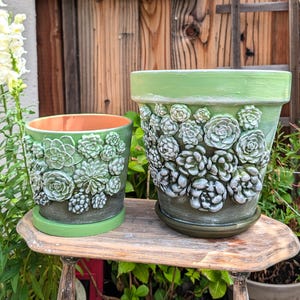 Op de afbeelding: Twee groene keramische bloempotten met verhoogde vetplantontwerpen. De grotere pot heeft een bijpassende schotel. De potten zijn geschilderd in groen- en wittinten en staan op een kleine houten tafel. De binnenkant van de kleinere pot is oranje.