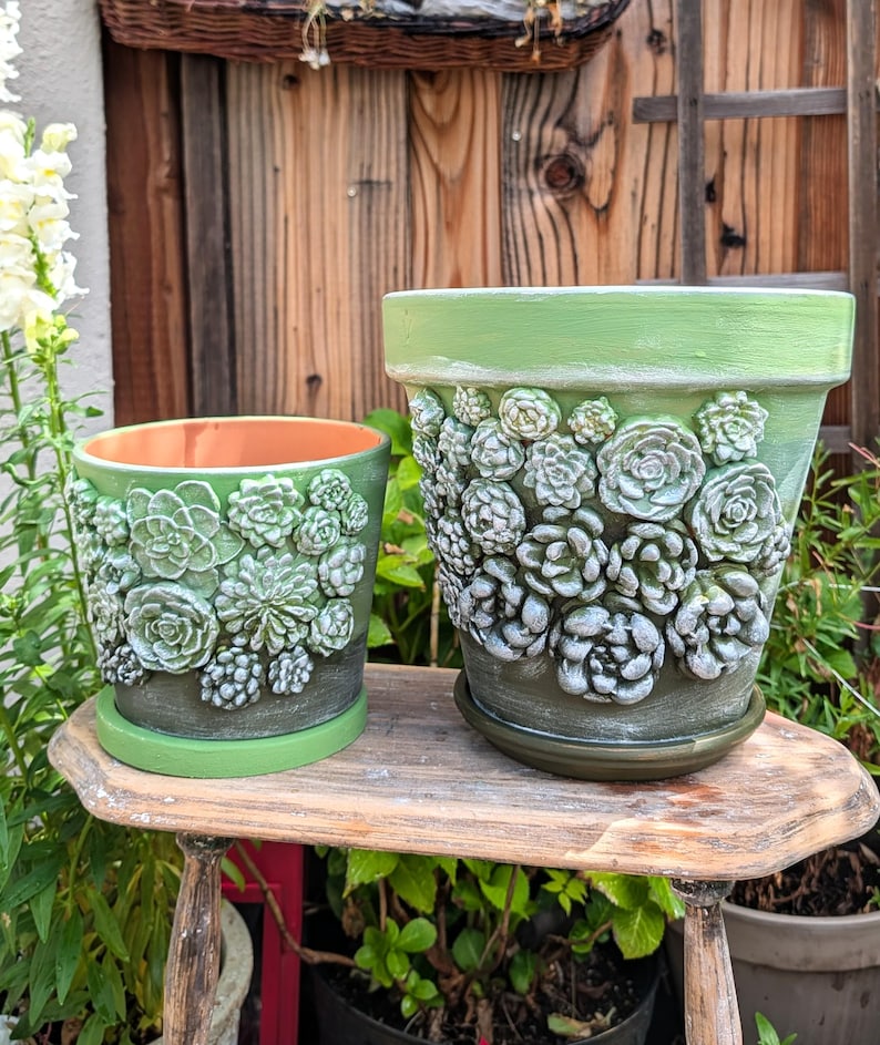 Op de afbeelding: Twee groene keramische bloempotten met verhoogde vetplantontwerpen. De grotere pot heeft een bijpassende schotel. De potten zijn geschilderd in groen- en wittinten en staan op een kleine houten tafel. De binnenkant van de kleinere pot is oranje.