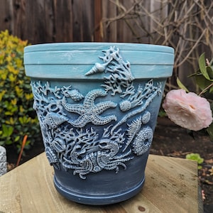 May include: A blue and white decorative flower pot with an ocean theme. The pot features embossed starfish, coral, and seashells. The pot has a gradient of blue hues, with a lighter blue at the top and a darker blue at the bottom.