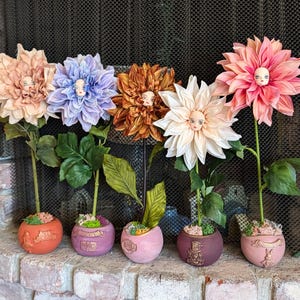 May include: Five artificial dahlia flowers in various colors, each with a face in the center. The flowers are in small, round pots with decorative designs. The pots are arranged on a brick surface.