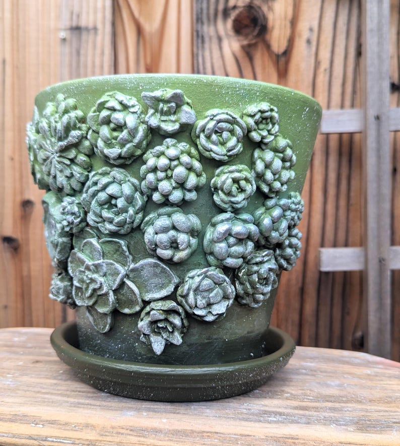 Op de afbeelding: Een groene keramische bloempot met bijpassende schotel. De pot is versierd met verhoogde, getextureerde succulentenontwerpen in groen- en zilvertinten. De pot staat op een houten oppervlak.