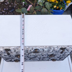Puede incluir: Caja de joyer&iacute;a de madera blanca con intrincados tallados florales y tres cajones. Cada caj&oacute;n tiene una perilla de lat&oacute;n. La caja se est&aacute; midiendo con una cinta m&eacute;trica.