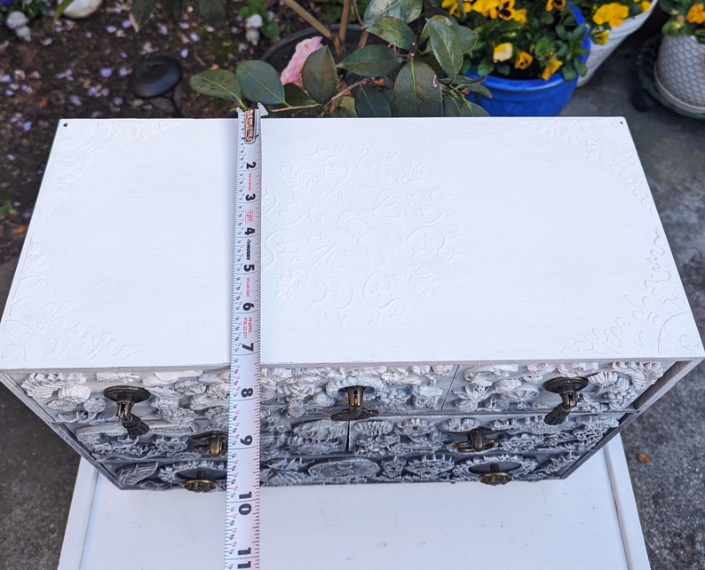 Puede incluir: Caja de joyer&iacute;a de madera blanca con intrincados tallados florales y tres cajones. Cada caj&oacute;n tiene una perilla de lat&oacute;n. La caja se est&aacute; midiendo con una cinta m&eacute;trica.