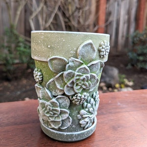Op de afbeelding: Een groene keramische bloempot met een getextureerde afwerking en witte spikkels. De pot is versierd met gedetailleerde, driedimensionale succulentenontwerpen. De pot staat op een houten oppervlak.