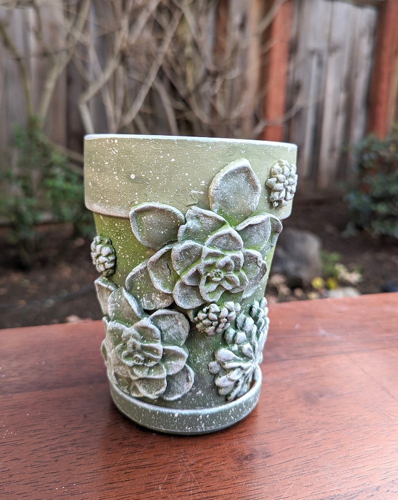 Op de afbeelding: Een groene keramische bloempot met een getextureerde afwerking en witte spikkels. De pot is versierd met gedetailleerde, driedimensionale succulentenontwerpen. De pot staat op een houten oppervlak.