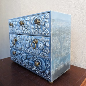 Puede incluir: Una c&oacute;moda de madera azul con tallas intrincadas y tiradores de metal decorativos. Cada caj&oacute;n est&aacute; adornado con un dise&ntilde;o &uacute;nico, con flores, hojas y otros motivos. La c&oacute;moda tiene un acabado desgastado, lo que le da un aspecto vintage.