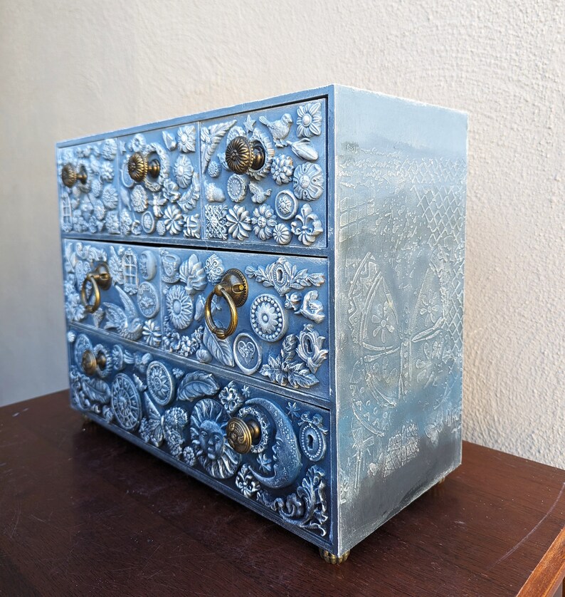 Puede incluir: Una c&oacute;moda de madera azul con tallas intrincadas y tiradores de metal decorativos. Cada caj&oacute;n est&aacute; adornado con un dise&ntilde;o &uacute;nico, con flores, hojas y otros motivos. La c&oacute;moda tiene un acabado desgastado, lo que le da un aspecto vintage.