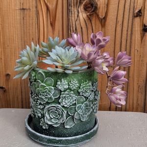 May include: A green ceramic planter with a detailed succulent design, filled with faux succulents and purple flowers. The planter sits on a matching saucer, with a wooden background.