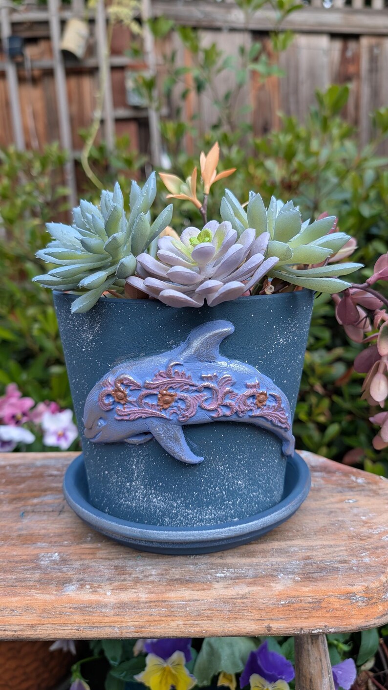 May include: A blue ceramic planter with a dolphin design and matching saucer, filled with succulents. The dolphin is light blue with ornate, reddish-brown accents. The planter sits on a wooden surface, with greenery in the background.