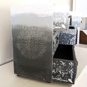 Puede incluir: Una peque&ntilde;a c&oacute;moda de madera gris con tres cajones. Los cajones tienen un dise&ntilde;o floral blanco y la c&oacute;moda tiene un acabado degradado blanco, gris y negro.
