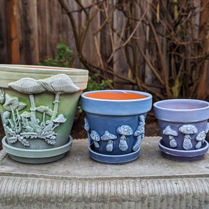 May include: Three ceramic flower pots with mushroom designs. The largest pot is green and white with a mushroom motif. The medium pot is blue and white with a mushroom motif. The smallest pot is purple and white with a mushroom motif.