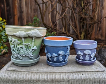 May include: Three ceramic flower pots with mushroom designs. The largest pot is green and white with a mushroom motif. The medium pot is blue and white with a mushroom motif. The smallest pot is purple and white with a mushroom motif.