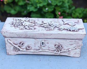 Può includere: Scatola rettangolare in legno bianco sporco con intagli floreali e di rami in rilievo. La scatola presenta un buco della serratura e una finitura strutturata e invecchiata. La parte superiore e anteriore sono decorate con dettagliati disegni botanici.