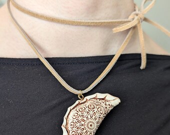 Può includere: Una collana girocollo in camoscio color cuoio con un ciondolo. Il ciondolo ha una forma a semicerchio con un design dettagliato e fantasia nei toni del marrone e crema. La collana ha una chiusura a fiocco.