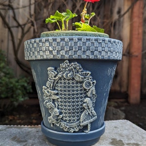 May include: A blue ceramic planter with a checkered pattern around the top rim. The planter features a raised design of Alice in Wonderland characters, including the Mad Hatter, the White Rabbit, and Alice. A small green plant with a single red flower is growing in the planter.