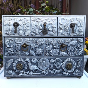 Puede incluir: Una caja de madera blanca con cuatro cajones. Cada caj&oacute;n est&aacute; decorado con un dise&ntilde;o diferente de detalles met&aacute;licos en relieve. Los dise&ntilde;os incluyen flores, hojas, p&aacute;jaros y otros elementos inspirados en la naturaleza.