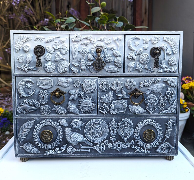Puede incluir: Una caja de madera blanca con cuatro cajones. Cada caj&oacute;n est&aacute; decorado con un dise&ntilde;o diferente de detalles met&aacute;licos en relieve. Los dise&ntilde;os incluyen flores, hojas, p&aacute;jaros y otros elementos inspirados en la naturaleza.