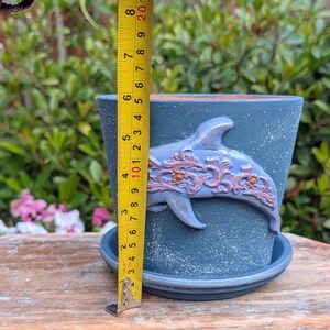 May include: A blue ceramic flower pot with a dolphin design. The pot is approximately 5 inches tall and has a matching saucer. The dolphin is light blue with decorative accents. The pot is on a wooden surface.