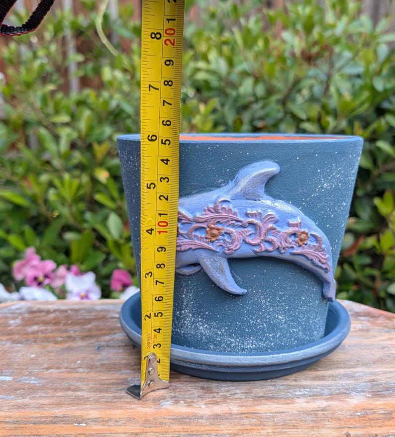 May include: A blue ceramic flower pot with a dolphin design. The pot is approximately 5 inches tall and has a matching saucer. The dolphin is light blue with decorative accents. The pot is on a wooden surface.