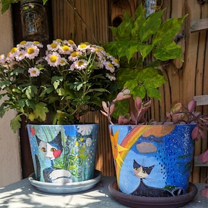 May include: Two hand-painted ceramic flower pots with cat designs. One pot features a cat with a red cheek, the other a cat under a rainbow. Both pots are blue and have matching saucers. The pots are filled with plants.