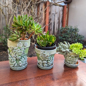 May include: A set of three ceramic mugs with a speckled, matte finish. The mugs are in shades of cream, light brown, and dark brown. Each mug has a simple, minimalist design and a rounded handle.