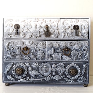 Puede incluir: Una caja de madera blanca y gris para joyas con intrincados tallados de flores, hojas y otros dise&ntilde;os inspirados en la naturaleza. La caja tiene cinco cajones con tiradores de lat&oacute;n y un acabado desgastado.