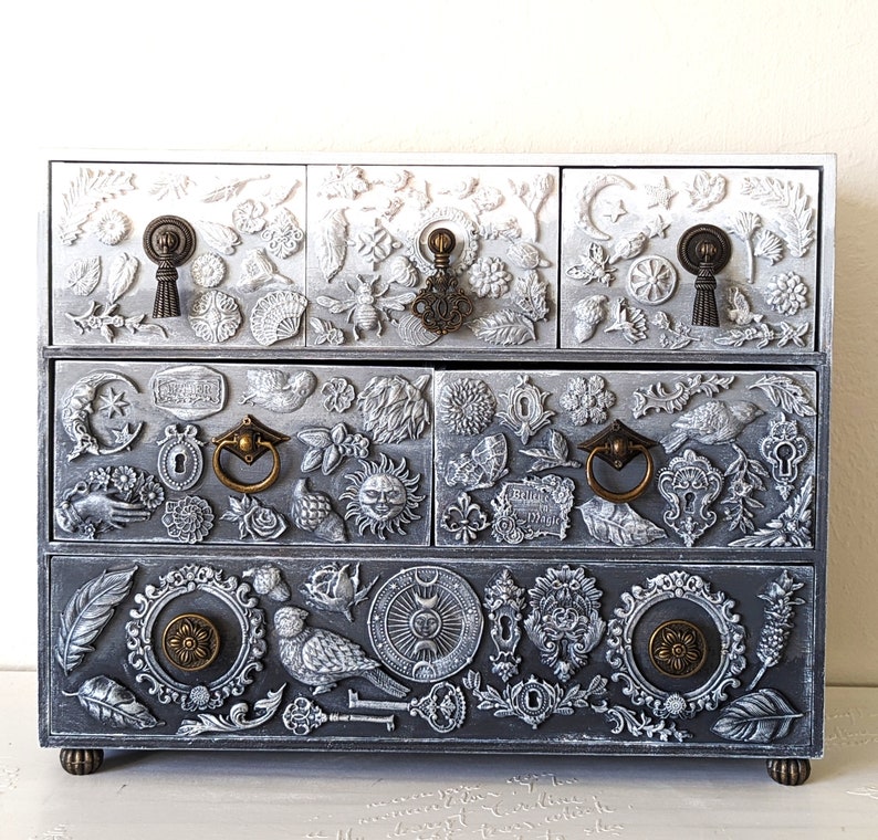 Puede incluir: Una caja de madera blanca y gris para joyas con intrincados tallados de flores, hojas y otros dise&ntilde;os inspirados en la naturaleza. La caja tiene cinco cajones con tiradores de lat&oacute;n y un acabado desgastado.