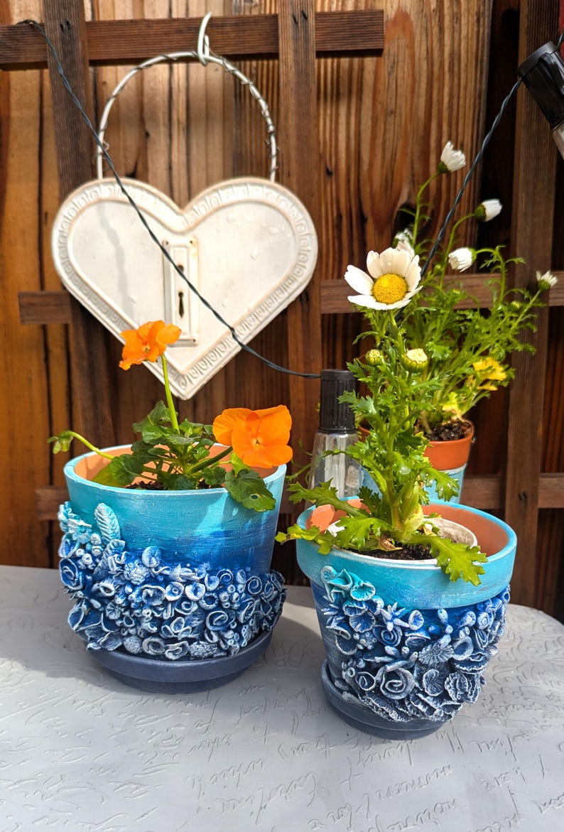 Pu&ograve; includere: Due vasi di fiori in ceramica blu e bianca con un design testurizzato a forma di conchiglia. Un vaso ha fiori arancioni e l'altro ha fiori bianchi.