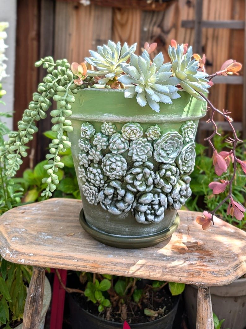 Op de afbeelding: Een groene keramische plantenpot met een gedetailleerd reli&euml;f van vetplanten. De pot is gevuld met levende vetplanten en hangend groen. De pot staat op een kleine houten kruk, wat een natuurlijke, aardse uitstraling cre&euml;ert.