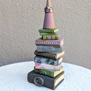 May include: A decorative stack of miniature books in various colors and textures, including green, purple, and grey, topped with a mauve cone-shaped ring holder. A metallic ring with a small stone is placed on the cone. The books feature embossed details, floral patterns, and a small oval frame on the bottom book.