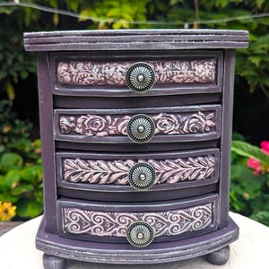 May include: A small, decorative jewelry box with four drawers. The box is painted a deep purple with silver accents. Each drawer features a floral design in pink and white, and a round, silver knob.