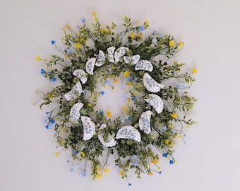 Può includere: Una ghirlanda circolare con una base di fogliame verde, accentuata da fiori gialli e blu. Pezzi di tessuto bianchi, semicircolari, con un motivo floreale blu sono distribuiti uniformemente.