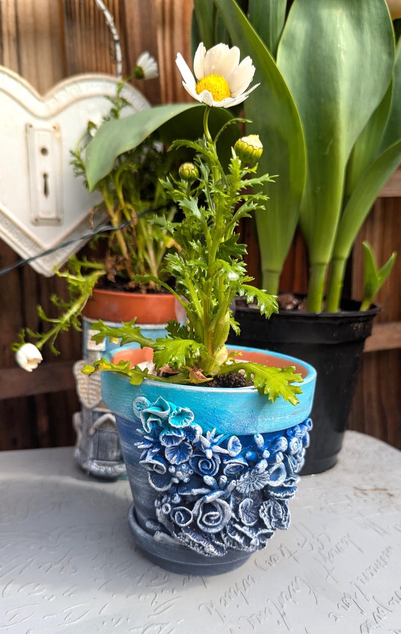 Pu&ograve; includere: Una margherita bianca con un centro giallo in un vaso di ceramica blu e bianco con un design decorativo. Il vaso &egrave; su una superficie bianca.