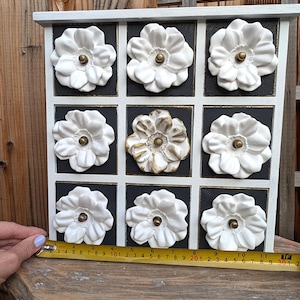 May include: A white and black decorative storage cabinet with nine square compartments. Each compartment features a white flower-shaped knob with a gold center. The cabinet is approximately 11 inches wide.