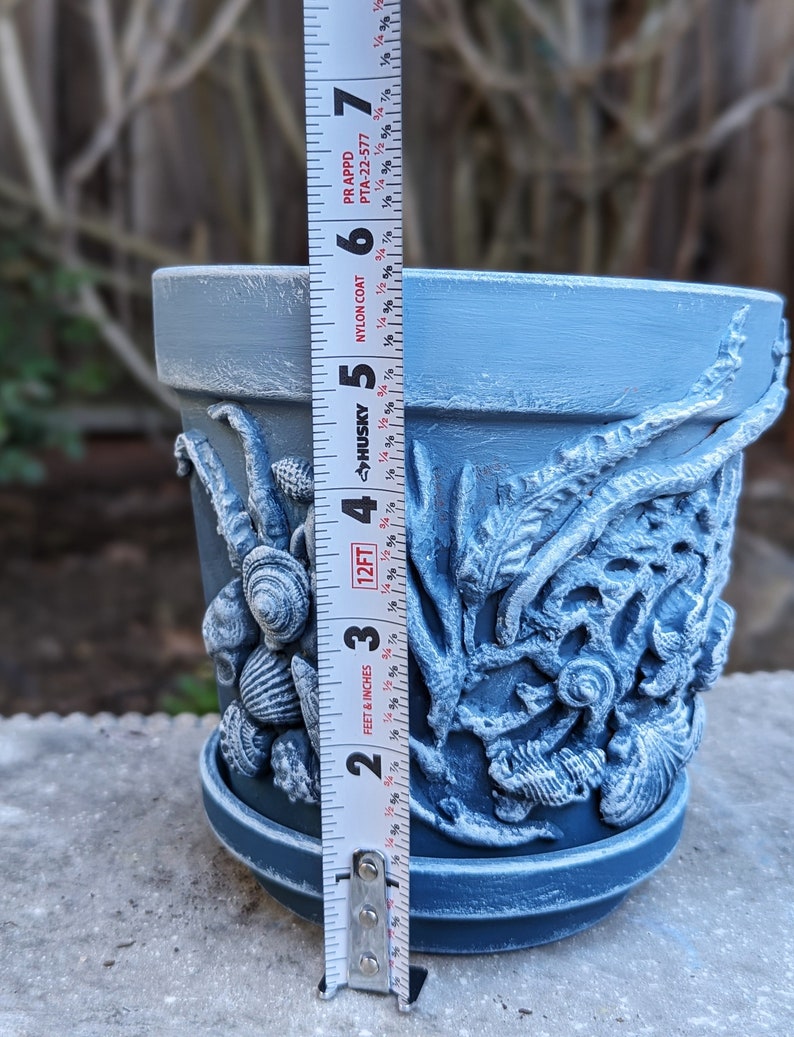May include: A blue painted ceramic flower pot with seashell and seaweed relief details. The pot is approximately 5 inches tall and includes a matching saucer. The pot is on a concrete surface.