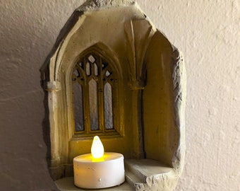 May include: A stone-colored candle holder shaped like a gothic window. The holder features a detailed window design and a small, battery-operated candle with a flickering yellow light. The candle sits on a small ledge within the window frame.