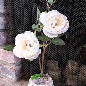 May include: A unique floral arrangement featuring white roses with doll faces in the center. The roses are on a brown stem with green leaves, placed in a white, geometric patterned pot. The arrangement sits in front of a black mesh screen.