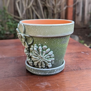 Op de afbeelding: Kleine groene keramische bloempot met bijpassende schotel. De pot is versierd met verhoogde, getextureerde vetplantontwerpen. De pot heeft een gespikkelde afwerking en een oranje interieur. Ideaal voor kleine planten.