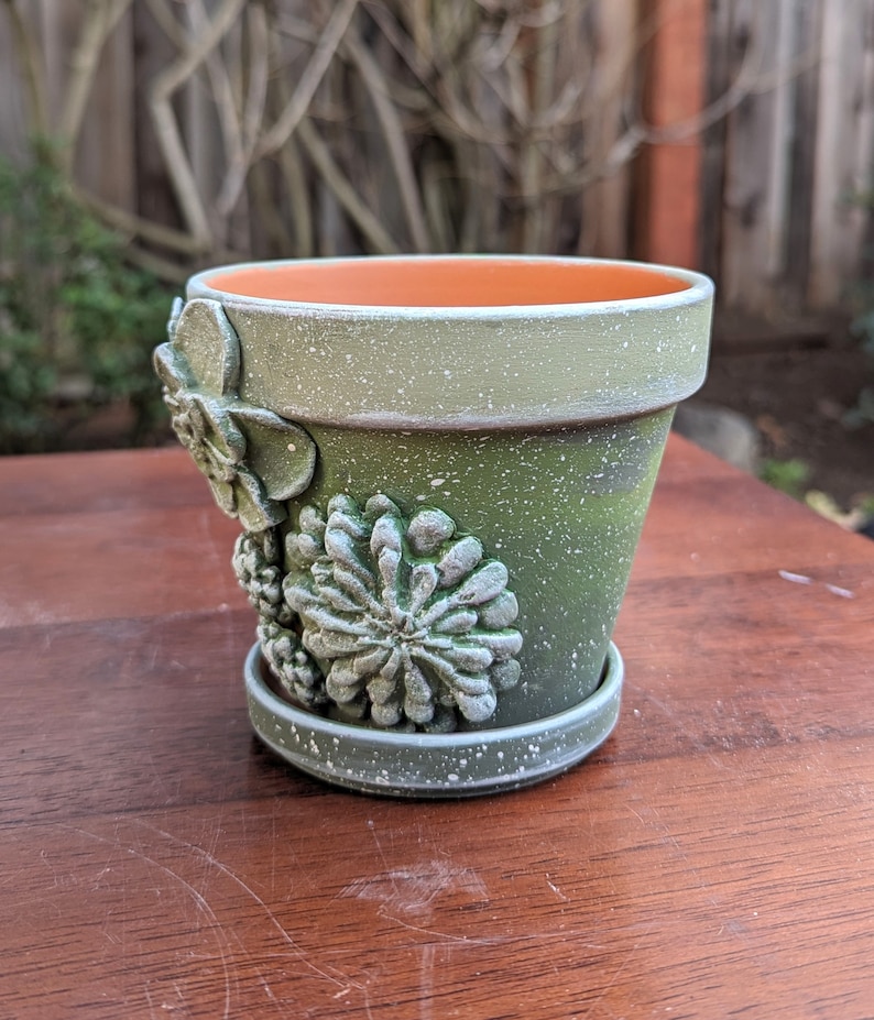 Op de afbeelding: Kleine groene keramische bloempot met bijpassende schotel. De pot is versierd met verhoogde, getextureerde vetplantontwerpen. De pot heeft een gespikkelde afwerking en een oranje interieur. Ideaal voor kleine planten.