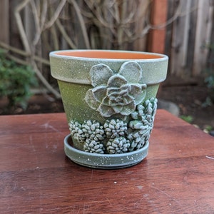 May include: A green ceramic plant pot with a matching saucer. The pot is decorated with a raised succulent design and speckled with white paint. The interior is orange.