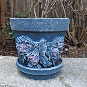 May include: A large, blue ceramic flower pot with a detailed relief of a bird with outstretched wings and floral accents. The pot has a textured surface and a matching saucer. The bird and flowers are in shades of blue and purple.