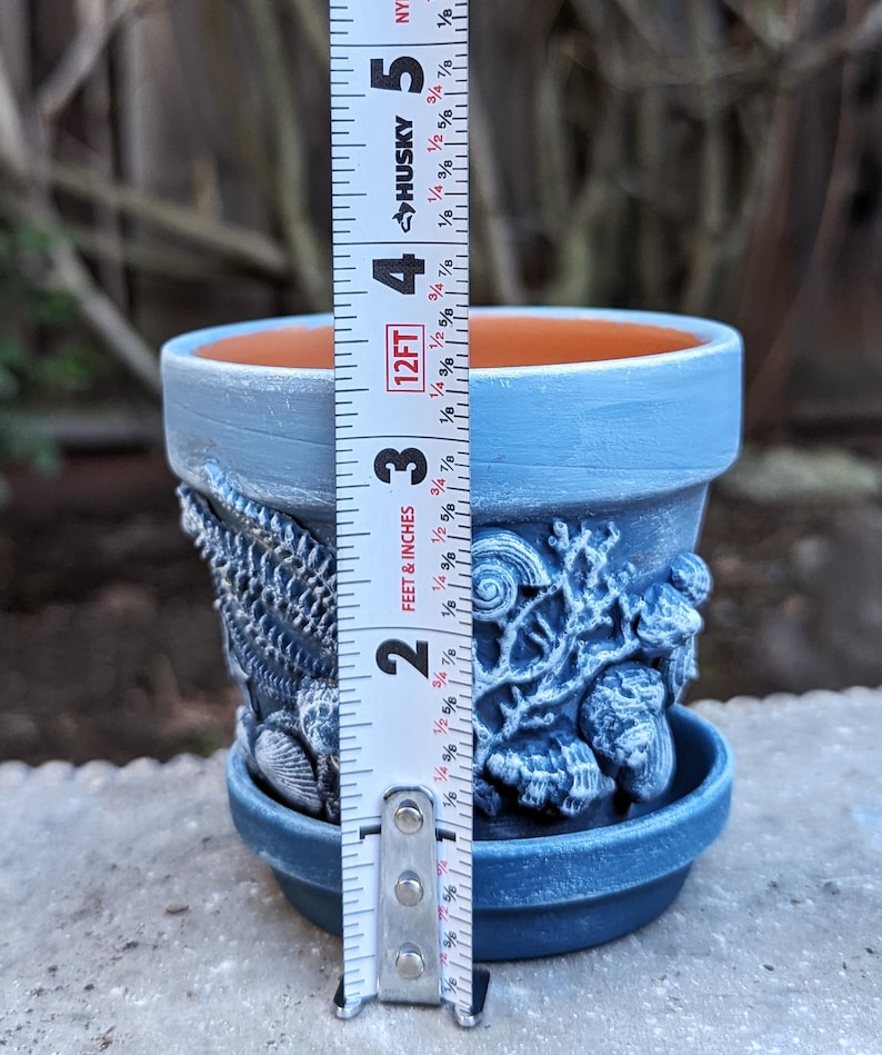 May include: A blue ceramic flower pot with a matching saucer. The pot is decorated with seashell and coral designs. The pot measures approximately 4 inches in height.