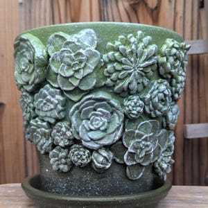 Op de afbeelding: Groene keramische plantenpot met bijpassende schotel. De pot is versierd met gedetailleerde, verhoogde succulentenontwerpen in verschillende maten. De pot heeft een gespikkelde afwerking en is geschikt voor binnen of buiten.