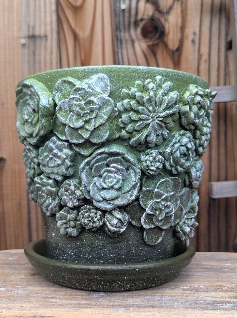 Op de afbeelding: Groene keramische plantenpot met bijpassende schotel. De pot is versierd met gedetailleerde, verhoogde succulentenontwerpen in verschillende maten. De pot heeft een gespikkelde afwerking en is geschikt voor binnen of buiten.