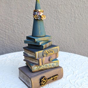 May include: A ceramic ring holder shaped like a stack of books with a cone on top. The cone is painted teal and gold and has several rings on it. The books are painted in shades of brown, gold, and teal. The bottom book has a gold key on it.